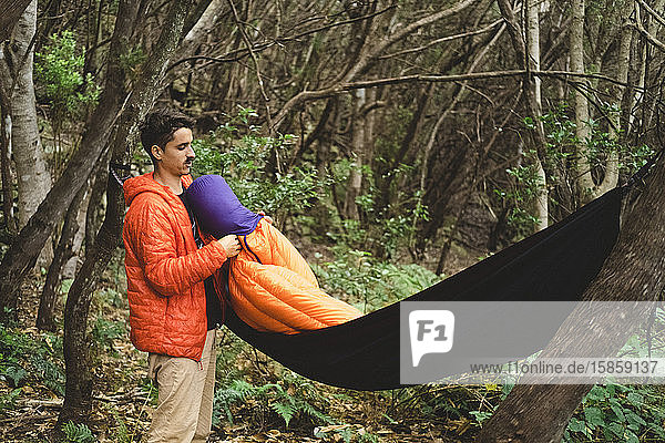 Ein Mann packt auf einem Campingplatz im Wald einen Schlafsack und eine Hängematte
