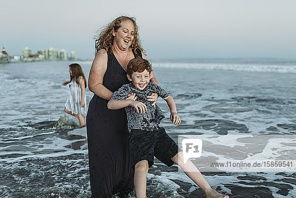 Young mother playing with young redhead son in ocean at dusk