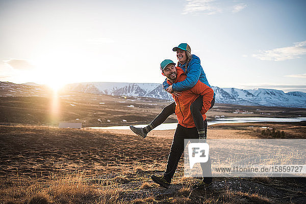 Man carrying woman on piggyback in field with mountains in background