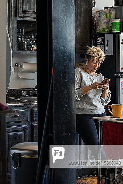 Lächelnde mittelgroße erwachsene Frau  die zu Hause in der Küche steht und Textnachrichten auf einem Smartphone verschickt