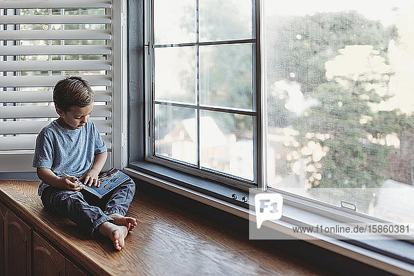 5 Jahre alter Junge liest ein Buch  während er auf einem Fensterplatz sitzt