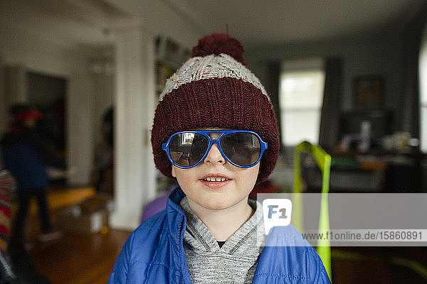 Mid shot of boy wearing blue sunglasses and winter hat indoors at home