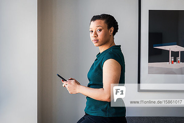 Seitenansicht der Textnachrichten einer Geschäftsfrau auf einem Smartphone  während sie im Büro an der Wand steht