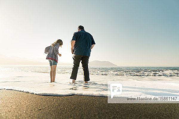 Father and daughter standing in water at beach during sunset