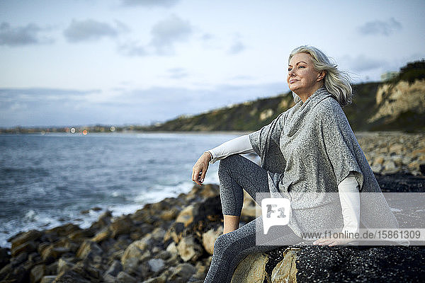 Seitenansicht einer nachdenklichen Frau  die bei Sonnenuntergang auf einem Felsen am Strand von Manhattan gegen den Himmel sitzt