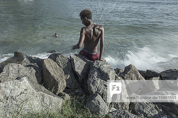 Wellenspritzer auf einen am Meeresufer auf Felsen sitzenden Mann