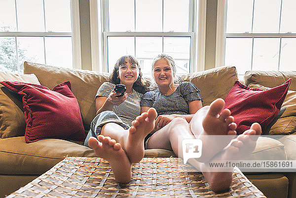 Two teenage girls sitting on couch with feet up watching television.