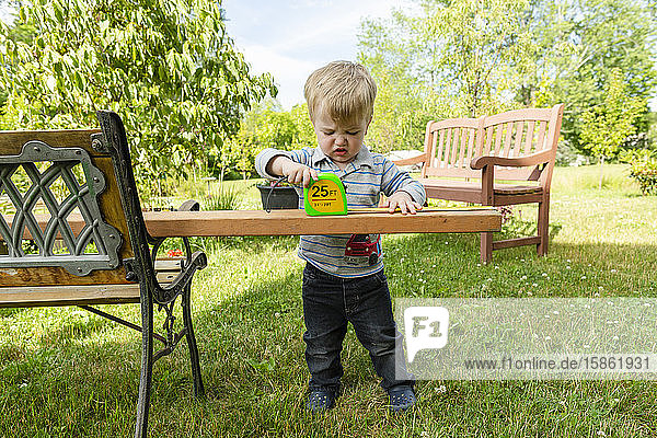 Kleinkind Junge sieht frustriert aus  während er im Freien ein Holzbrett misst