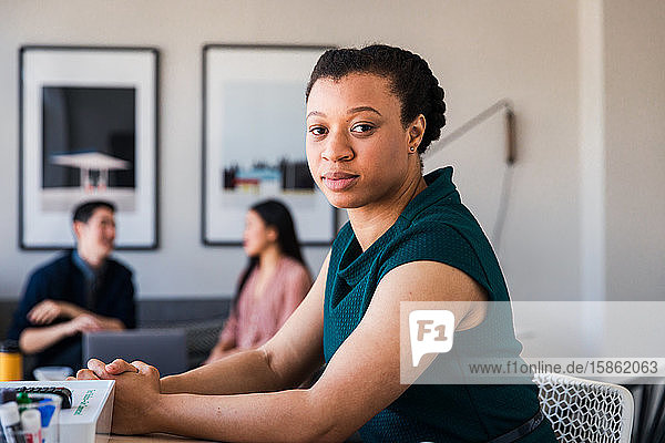 Porträt einer selbstbewussten Geschäftsfrau  die mit Kollegen im Hintergrund am Tisch sitzt