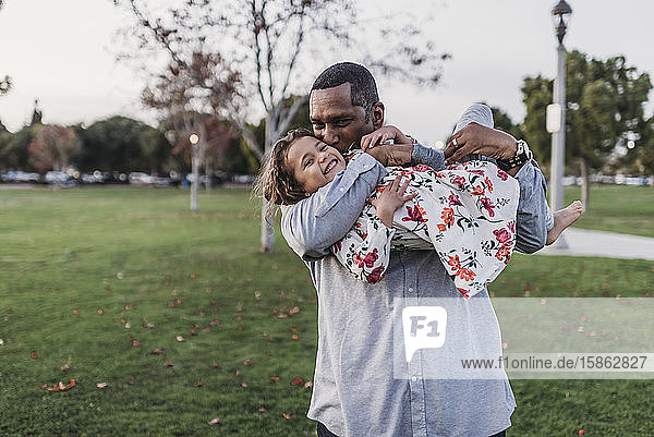 Glücklicher Vater hält süßes Mädchen in der Abenddämmerung auf einem Spielplatz im Park