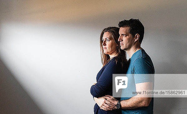 Couple standing close together in a patch of light against white wall.