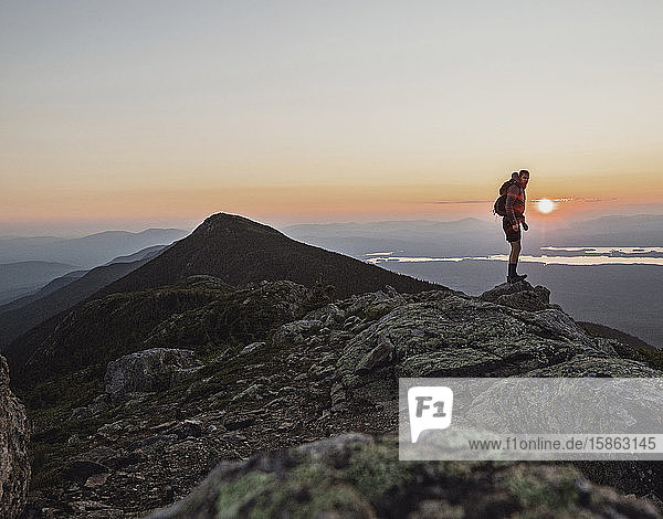 A male hiker stands at the summit of a mountain at sunset  Maine.