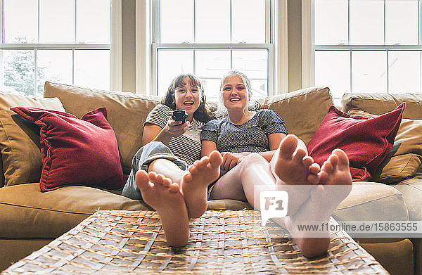 Two teenage girls sitting on a couch with feet up watching television.