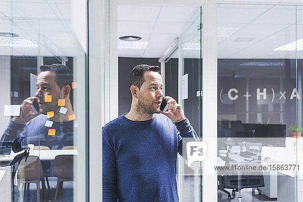 Seitenansicht eines Geschäftsmannes  der mit einem Smartphone spricht  während er an einer Bürowand steht