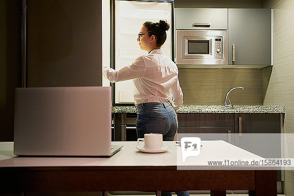 Eine junge Frau  die mit einem Laptop arbeitet  schaut in den Kühlschrank