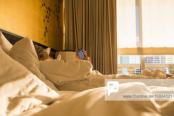 Erwachsene weibliche Führungskraft liegt in der frühen Morgensonne auf Hotelbett und schaut auf Smartphone