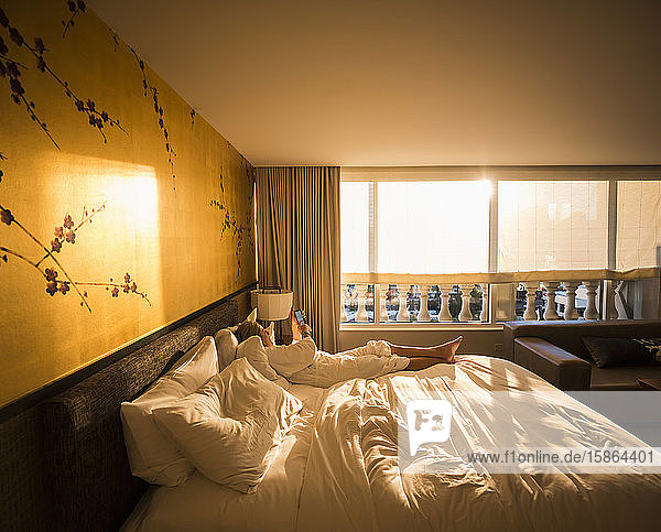 Erwachsene weibliche Führungskraft liegt in der frühen Morgensonne auf Hotelbett und schaut auf Smartphone