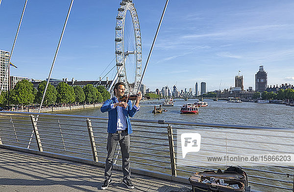 Busker playing violin on a bridge over the River Thames with boats and the London Eye in the background; London England