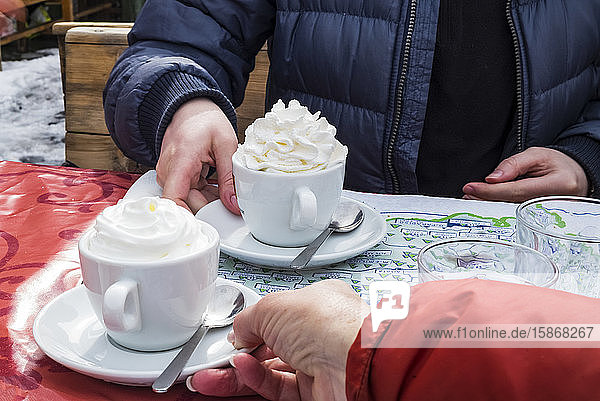 Ein Paar in Skijacken sitzt an einem Tisch und bekommt Tassen mit heißer Schokolade und Schlagsahne serviert; Courmayeur  Aostatal  Italien