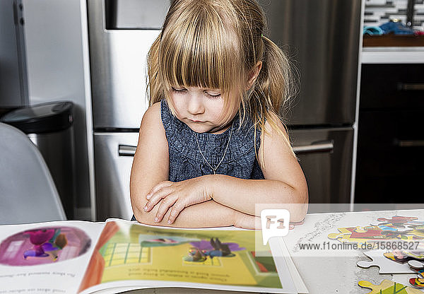 Ein süßes kleines Mädchen  das ein Bilderbuch liest und ein Puzzle auf einem Tisch in der Küche macht: Edmonton  Alberta  Kanada