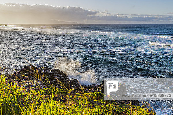 Ozeanwellen  die bei Sonnenuntergang an das felsige und grasbewachsene Ufer rollen  mit einer ruhigen Meereslandschaft und Wolken am Horizont des Ho'okipa Lookout bei Paia; Maui  Hawaii  Vereinigte Staaten von Amerika