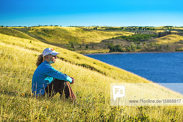 Frau sitzt auf einem grasbewachsenen Hügel mit Blick auf einen See und sanfte Hügel im Hintergrund mit blauem Himmel  Waterton Lakes National Park; Waterton  Alberta  Kanada