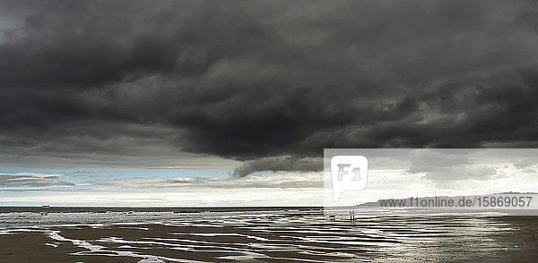 Dunkle Sturmwolken über dem Atlantischen Ozean mit zwei Menschen und ihrem Hund  die am nassen Sandstrand im Vordergrund spazieren gehen; South Shields  Tyne and Wear  England