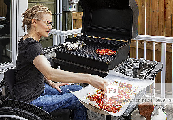 Eine querschnittsgelähmte Frau  die in ihrem Rollstuhl sitzend Steaks und Bratkartoffeln auf einem Grill im Freien zubereitet: Edmonton  Alberta  Kanada