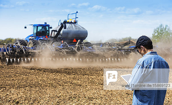 Farmer using a tablet while standing on a farm field and a tractor and equipment seeds the field; Alberta  Canada