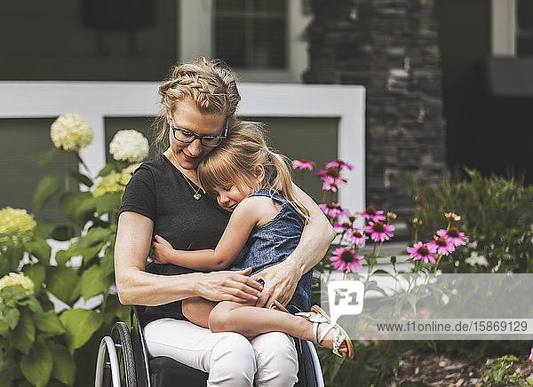 Eine querschnittsgelähmte Mutter hält ihr kleines Mädchen auf dem Schoß  während sie an einem warmen Sommernachmittag in ihrem Vorgarten im Rollstuhl sitzt: Edmonton  Alberta  Kanada.