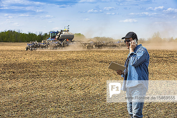 Farmer using a smart phone and tablet while standing on a farm field and watching the tractor and equipment seeding the field; Alberta  Canada