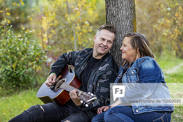 A mature couple spending quality time together and the wife is listening to her husband singing and playing his guitar while in a city park on a warm fall evening: Edmonton  Alberta  Canada
