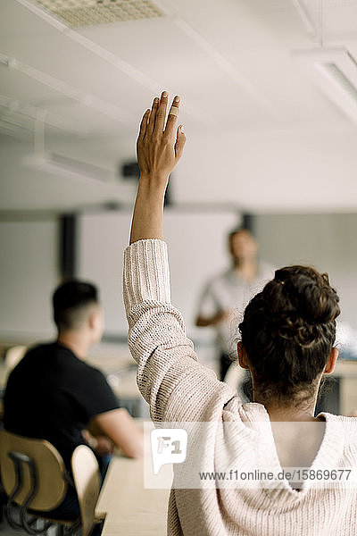 Female student with hand raised while tutor standing in classroom
