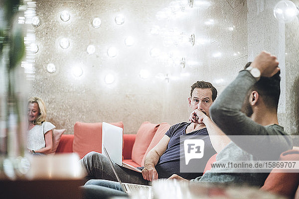 Geschäftsmann sitzt mit einem Kollegen zusammen  während er seinen Laptop auf dem Sofa im Kreativbüro benutzt