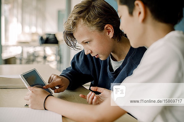 Männlicher Student zeigt auf digitales Tablett  während er mit einem Freund im Klassenzimmer sitzt