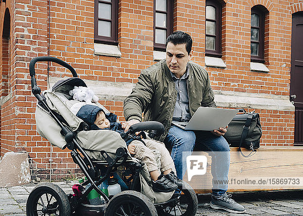 Vater benutzt Laptop  während der Sohn im Kinderwagen gegen Gebäude in der Stadt sitzt