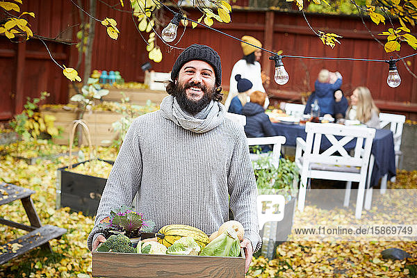 Porträt eines glücklichen Mannes mit Gemüsekorb stehend  während Familie und Freunde am Tisch im Hof sitzen
