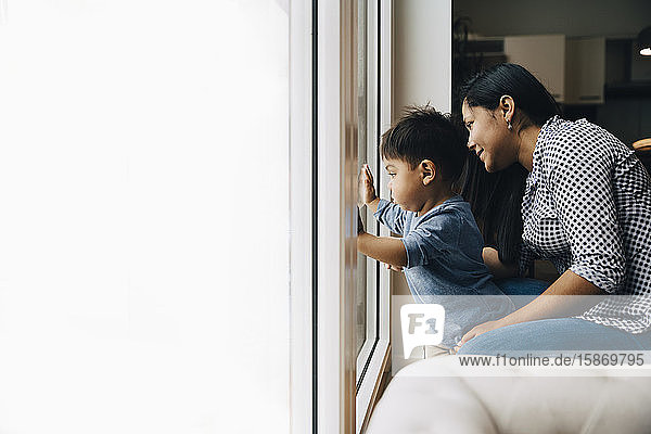 Side view of mother and son looking through window while sitting at home