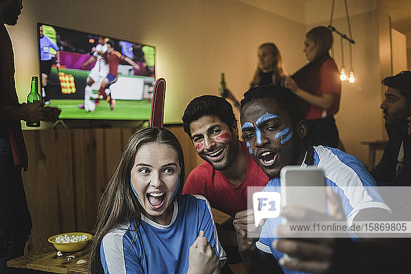 Fans nehmen Selfie mit dem Handy  während Freunde ein Fußballspiel im Fernsehen anschauen