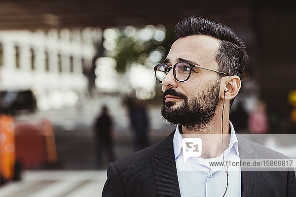 Confident businessman with in-ear headphones looking away while standing outdoors