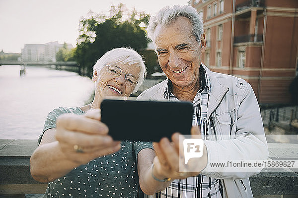 Lächelndes älteres Pärchen  das sich mit dem Handy selbstständig macht  während es in der Stadt am Geländer steht