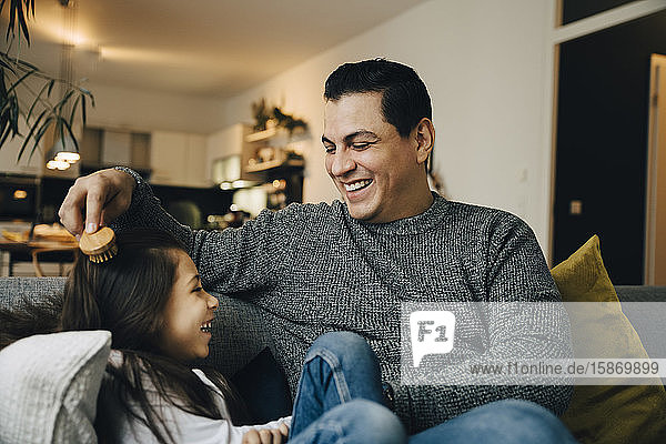 Smiling father combing daughter's hair while sitting on sofa at home