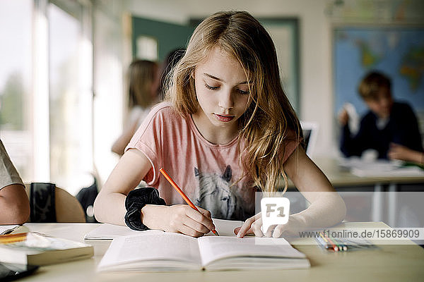 Studentin schreibt in Buch  während sie im Klassenzimmer am Tisch sitzt