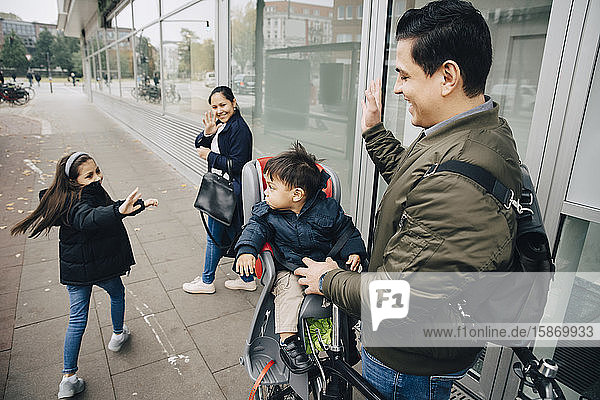 Lächelnde Mutter und Tochter winken dem Jungen mit dem Vater auf dem Bürgersteig in der Stadt