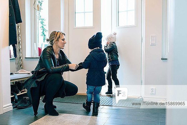Mother helping daughter in wearing jacket while crouching at home