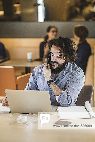 Männlicher Unternehmer mit Laptop  der mit seinem Handy telefoniert  während er im Büro am Tisch sitzt