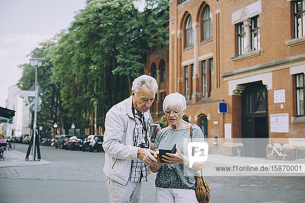 Ältere Frau hält Smartphone in der Hand und diskutiert mit Partner während einer Erkundungstour in der Stadt