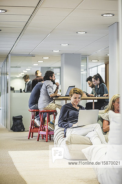 Geschäftsfrau benutzt Laptop  während Kollegen im Hintergrund im Büro arbeiten