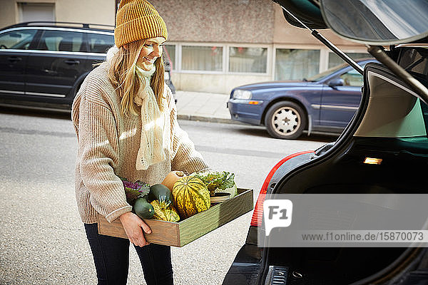 Glückliche Frau mit frisch produziertem Gemüse in einem Container  der neben dem Kofferraum eines Autos steht