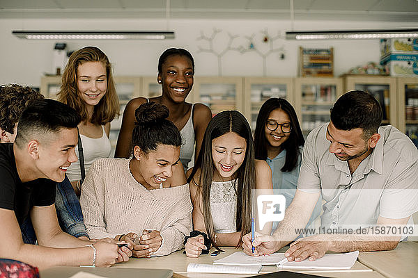 Lächelnder Tutor unterrichtet männliche und weibliche Studenten  während er sich im Klassenzimmer über den Tisch lehnt